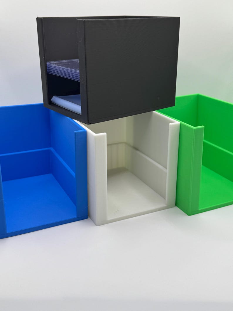 May include: A black, rectangular card storage box with a stack of cards inside, hovering above three open-top boxes in blue, white, and green. The boxes are designed for organizing and storing cards.