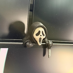 Può includere: Una piccola figura con cappuccio nero e una maschera bianca allungata con un'espressione a bocca aperta. La figura ha maniche nere e tiene in mano un piccolo coltello color argento, sporgendosi oltre un bordo sottile e scuro.