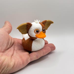 Może przedstawiać: Figurka gumowej kaczki inspirowana Gremlinami. Kaczka ma brązowe, białe i pomarańczowe kolory, duże uszy i psotny wyraz twarzy. Kaczka jest trzymana w dłoni na białym tle.