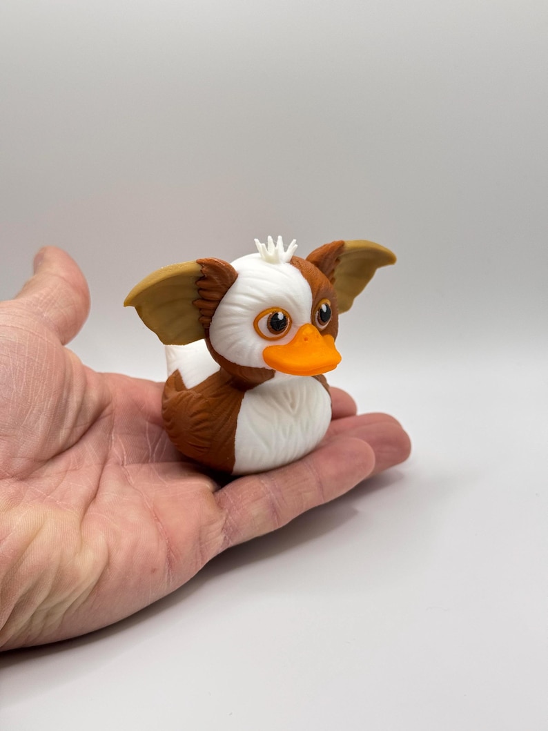 Może przedstawiać: Figurka gumowej kaczki inspirowana Gremlinami. Kaczka ma brązowe, białe i pomarańczowe kolory, duże uszy i psotny wyraz twarzy. Kaczka jest trzymana w dłoni na białym tle.