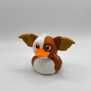 Może przedstawiać: Nowatorska gumowa zabawka w kształcie Gremlina. Kaczka ma brązowe, białe i pomarańczowe kolory, z dużymi uszami i psotnym wyrazem twarzy. Unikalny przedmiot kolekcjonerski.