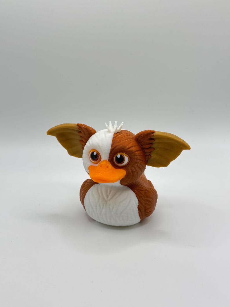 Może przedstawiać: Nowatorska gumowa zabawka w kształcie Gremlina. Kaczka ma brązowe, białe i pomarańczowe kolory, z dużymi uszami i psotnym wyrazem twarzy. Unikalny przedmiot kolekcjonerski.