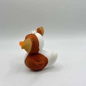 Może przedstawiać: Kapryśna gumowa zabawka w kształcie kaczki z brązowym korpusem, białymi akcentami i pomarańczowym dziobem. Kaczka ma brązowe uszy i małą białą koronę. Zabawka została zaprojektowana tak, aby przypominać kaczkę.