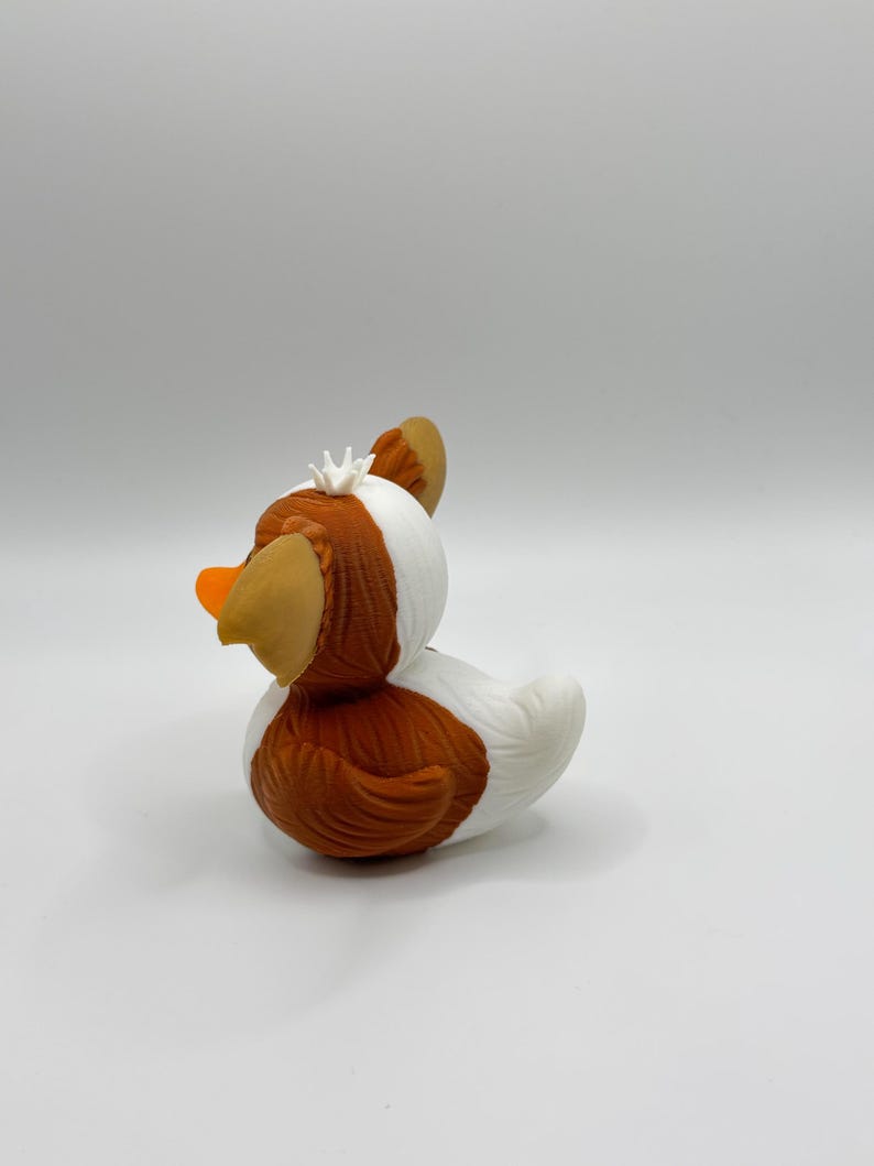 Może przedstawiać: Kapryśna gumowa zabawka w kształcie kaczki z brązowym korpusem, białymi akcentami i pomarańczowym dziobem. Kaczka ma brązowe uszy i małą białą koronę. Zabawka została zaprojektowana tak, aby przypominać kaczkę.