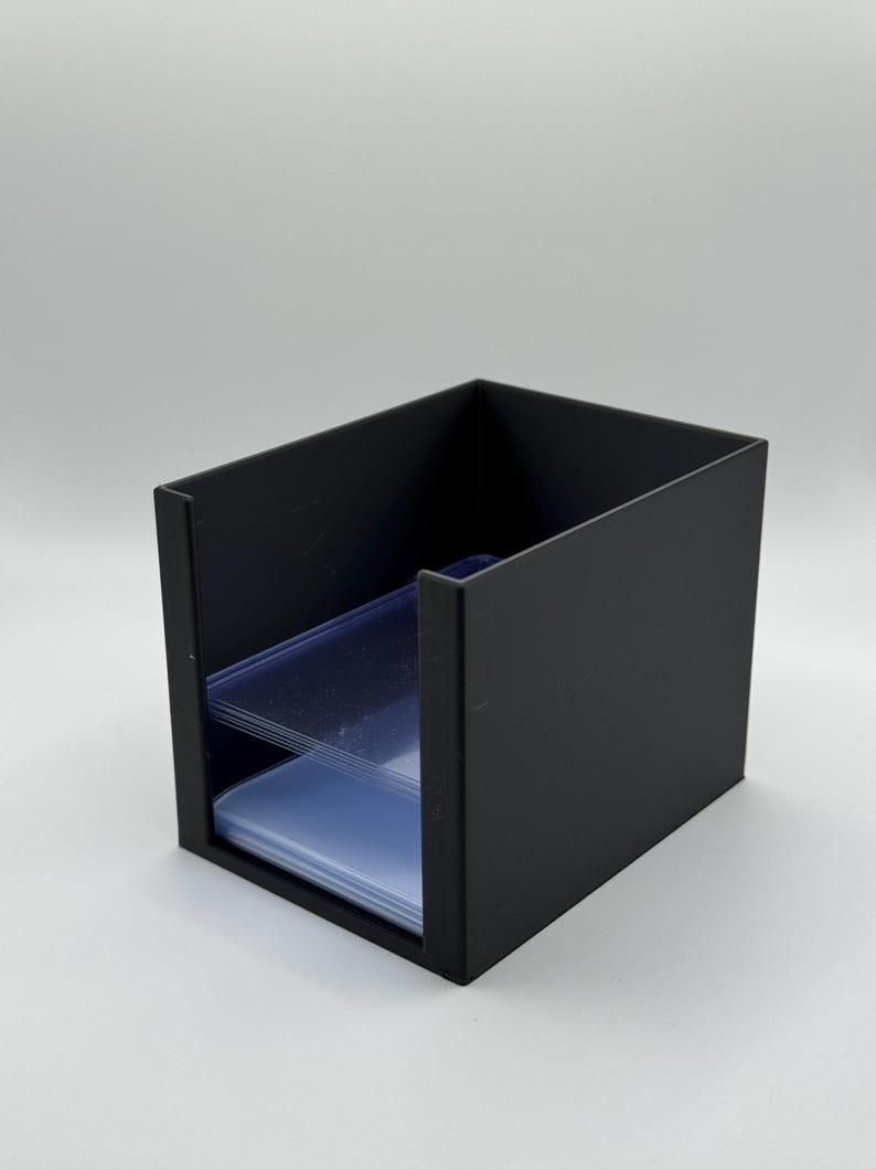 May include: A black, rectangular card holder with two shelves. The holder is made of a solid, matte black material. Clear, plastic card sleeves are visible on the shelves. The holder is designed to store and organize trading cards or similar items.