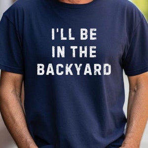 May include: A navy blue t-shirt with white text that reads "I'll be in the backyard".