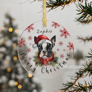 May include: Clear, round Christmas ornament featuring a Boston Terrier wearing a Santa hat and scarf. The ornament has the name "Sophie" and the year "2024" printed on it, with "Merry Christmas" text and red snowflake accents.