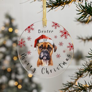 May include: Round glass Christmas ornament with a red and white snowflake pattern. A brown and white boxer dog wearing a red Santa hat is in the center of the ornament. The text "Dexter" and "Merry Christmas" are on the ornament.