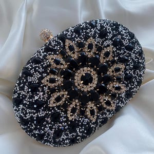 May include: A black and silver oval clutch bag with a large black rhinestone embellishment in the centre. The bag is covered in small rhinestones.