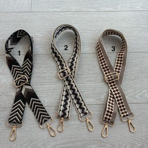 May include: Three patterned bag straps with gold-coloured hardware. The straps feature different designs: a black and white chevron, a black and grey zigzag, and a brown and white checkered pattern. Each strap has a metal clasp.
