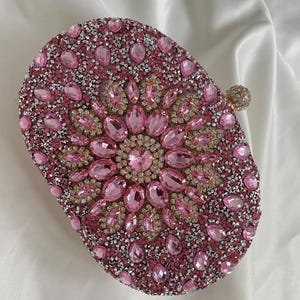 May include: A pink oval-shaped clutch purse covered in pink and silver rhinestones. The centre of the purse features a large pink rhinestone flower.