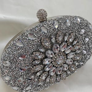 May include: A silver clutch bag covered in sparkling rhinestones and crystals. The bag has a round, silver clasp.