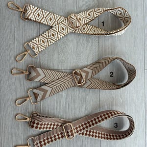 May include: Three decorative bag straps with gold-coloured hardware. The straps feature woven patterns in cream, brown, and tan. Designs include geometric diamond, chevron, and checkered patterns. Each strap has a gold-coloured metal buckle and two gold-coloured metal clasps.