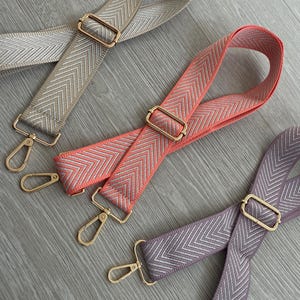 May include: Three adjustable bag straps in beige, coral, and lavender. Each strap has a chevron pattern and gold-coloured metal hardware, including rectangular buckles and swivel clasps. The straps are displayed on a light grey wooden surface.
