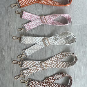 May include: Five patterned bag straps in shades of orange, pink, cream, beige, and brown. Each strap has a geometric design and gold-coloured metal hardware, including clasps and adjustable buckles. The straps are displayed on a light wood-grain surface.