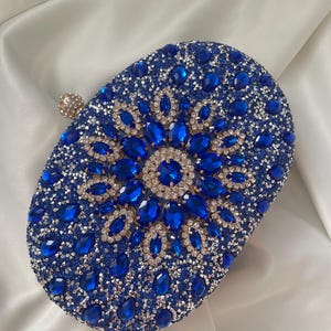 May include: An oval-shaped clutch purse covered in rhinestones and crystals. The purse is blue and silver with a large blue crystal design in the centre.