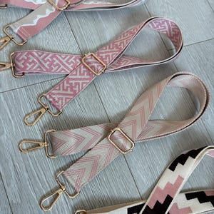 May include: Four patterned bag straps in different designs and colours. The straps feature pink, white, and black geometric patterns, with gold-coloured metal hardware. The straps are designed to be interchangeable and add a decorative element to a bag.