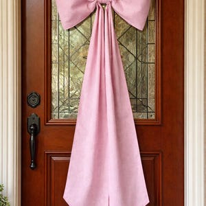 May include: A pink fabric door bow with a long, draping tail hangs on a wooden door with a glass window. The bow is made of textured fabric and is a soft, pastel pink color. The door has a dark handle.