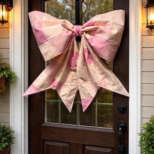 May include: A large, pink quilted bow hangs on a dark brown door. The bow is made of various pink and cream-colored fabrics, creating a patchwork design. The door has a glass window and black hardware, with a light fixture on either side.
