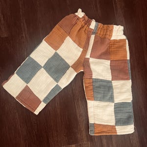 Puede incluir: Pantalones de bebé con diseño de patchwork. Los pantalones presentan cuadrados blancos, azules, marrones y naranjas. La cintura es elástica. La tela es suave y ligera.