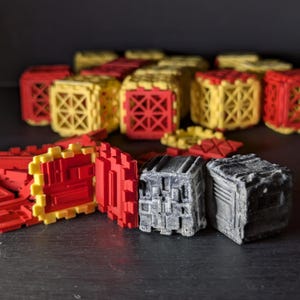 May include: A close-up of two grey, textured, cube-shaped blocks. The blocks are sitting on a black surface. Behind the blocks are several red and yellow plastic blocks.
