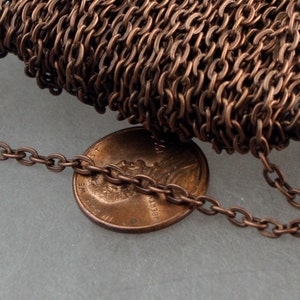 May include: A spool of antique copper-colored metal cable chain with small oval links. The chain is positioned next to a United States one-cent penny to provide a visual scale reference for the size of the individual chain links.
