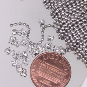 May include: A silver-toned ball chain and several matching metal connectors are displayed next to a copper-colored United States one-cent coin for scale. The chain consists of small, uniform spherical beads linked together in a continuous strand.