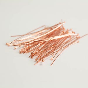 Peut inclure: Une collection d'épingles à tête en or rose, chacune composée d'une tige métallique fine et droite surmontée d'une petite boule sphérique. Ces composants de bijouterie sont disposés en un tas lâche et superposé, révélant leur fini métallique.