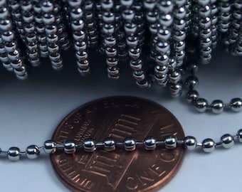 May include: A coiled length of silver-toned metal ball chain featuring small, uniform spherical beads connected by short links. The chain is positioned in front of a copper-colored United States one-cent coin, which provides a scale reference.
