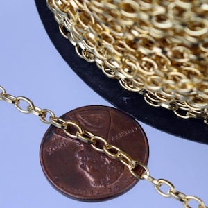 May include: Close-up of a gold-colored oval link chain. A penny is next to the chain for size comparison. The chain is coiled on a black spool. The chain links are small and closely connected, creating a delicate and shiny appearance.