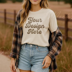 Puede incluir: Una persona lleva una camiseta color crema con el texto "Your Design Here" en escritura marrón. También lleva pantalones cortos de mezclilla y una camisa de franela a cuadros marrón. El fondo es una escena borrosa al aire libre.