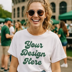May include: A white t-shirt with the text "Your Design Here" in green, worn by a person with sunglasses and a ponytail. The person is smiling in front of a blurred outdoor background.