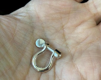May include: A small, shiny silver shackle with a screw closure. The shackle is in the shape of a U, with a rounded top and a threaded screw on one end. The screw has a small, flat head.