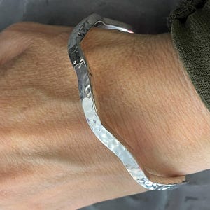 May include: A hammered silver bracelet with a wavy design. The bracelet has a textured surface and is worn on a wrist. The metal reflects light, creating highlights and shadows. The bracelet is a piece of jewelry.