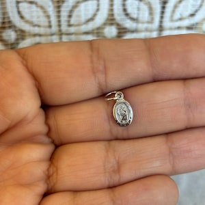 May include: A small, silver-colored pendant with an oval shape. The pendant features a detailed image of a religious figure. The pendant is held in a person's hand, showcasing its size and design. The pendant has a small loop for a chain.