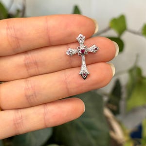 May include: A silver cross pendant with a red gemstone in the centre and smaller red gemstones around the edges. The cross is decorated with small white gemstones.