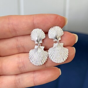 May include: Silver tone double seashell dangle earrings. The earrings feature two textured seashells, one smaller and one larger, connected by a small loop.