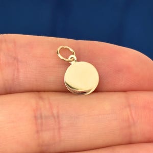 May include: A small, round gold charm with a loop for attaching to a necklace or bracelet. The charm is a solid, polished gold color and has a smooth surface. The charm is held in a person's hand against a blue background.