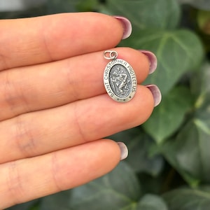 May include: Silver oval pendant with a depiction of Saint Christopher carrying a child. The pendant has the text "Saint Christopher Protect Us" around the edge.