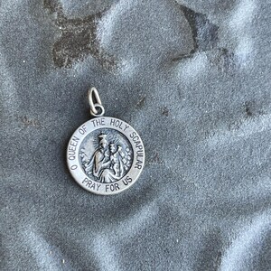 May include: A round silver-toned metal pendant featuring a raised relief image of a figure holding a child. The text O QUEEN OF THE HOLY SCAPULAR PRAY FOR US is embossed around the circular border. A small silver-toned jump ring is attached to the top.