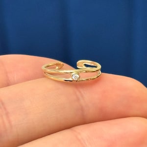 May include: A gold-colored double-band ring with a small, clear gemstone. The ring is set against a blue background. The ring is a delicate piece of jewelry.