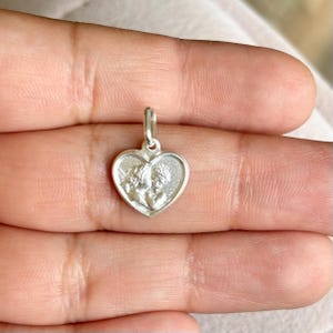 Op de afbeelding: Een zilveren hartvormige hanger met een gedetailleerd reli&euml;f van twee cherubs. De hanger heeft een kleine lus aan de bovenkant voor een ketting. De cherubs kijken elkaar aan. De hanger wordt in een hand gehouden.