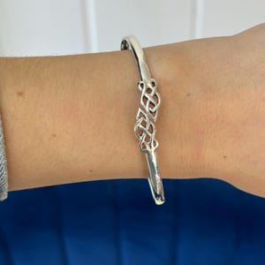 May include: A silver bangle bracelet with a Celtic knot design. The bracelet is open and has a simple, elegant look.