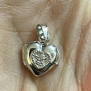 May include: A silver heart-shaped locket pendant. The locket has a small heart design on the front and a loop for a chain. The locket is shiny and reflective.