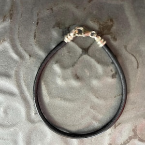 Puede incluir: Una pulsera de cuero negro con un cierre plateado. La pulsera es circular y parece estar hecha de un material fino y flexible. El cierre es de tipo mosquetón. La pulsera se muestra sobre un fondo gris texturizado.