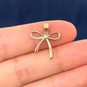 May include: A gold-colored bow pendant. The pendant is in the shape of a bow with two loops and two flowing tails. The pendant is small and delicate, with a simple design. The background is a solid dark blue.