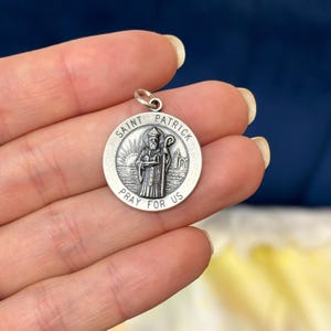 May include: A silver Saint Patrick pendant with a detailed image of Saint Patrick holding a staff, with the text "Saint Patrick" and "Pray for Us" around the edge. The pendant is round and has a small loop at the top.