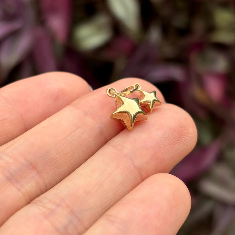 May include: Two gold star charms, one slightly larger than the other, are shown in a hand. The charms are simple and elegant, with a smooth, polished finish.