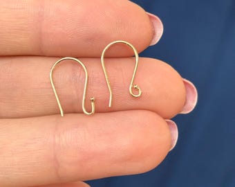May include: Two gold-colored earring hooks are displayed. The hooks are in a curved, open-loop design, with a small ball at the bottom of one hook. The hooks are held between fingers against a blue background.