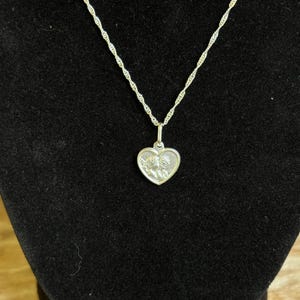 Op de afbeelding: Een zilverkleurige ketting met een hartvormige hanger. De hanger heeft een gedetailleerd reli&euml;fontwerp en hangt aan een delicate ketting. De ketting wordt gepresenteerd tegen een zwarte achtergrond.